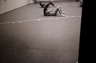 Two women in athletic wear perform stretching exercises on a polished indoor surface, possibly a gymnasium or studio. The spa...