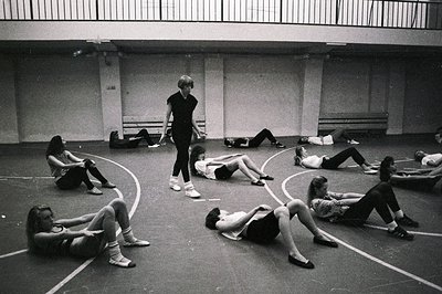 A ballet class in progress; a female instructor, center, directs a group of female students in a floor exercise. The dancers ...