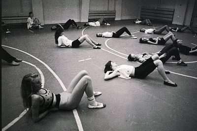 A black and white image captures a dance class in progress. Numerous students, likely ballet or modern dancers, are positione...