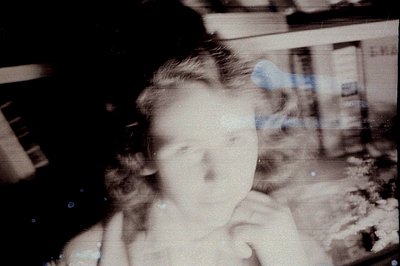 A faded, slightly blurred color photograph depicts a young girl with short, dark hair. The image exhibits significant film gr...