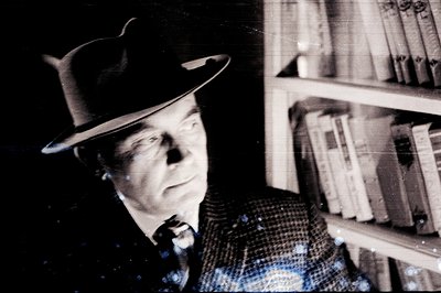 A man in a fedora and patterned jacket looks off-camera, framed by a tall bookshelf. The black and white photograph has visib...