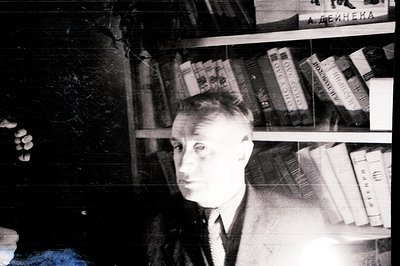 A black and white portrait depicts a man in a suit seated amongst bookshelves. Cyrillic script is visible on the book spines....