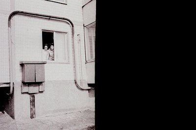 A young woman peeks out from a window within a concrete courtyard structure. Exposed utility pipes and an electrical box are ...