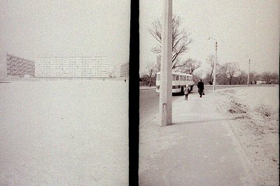 A monochrome street scene featuring a brutalist apartment block, bus, and two figures bundled in winter clothing. Likely East...