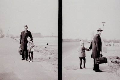 A man in a dark coat and hat holds a child's hand as they walk on a snow-covered street. The background reveals large, modern...