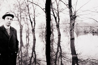 A man in a dark coat and hat stands formally posed. Sparse trees and a building visible in the background suggest a rural, wi...