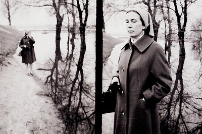 A woman in a dark coat & headscarf stands formally on a slight incline, a figure visible in the distance. The composition emp...