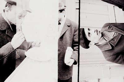 Black and white image shows two individuals viewed through a window pane. A man in a coat and hat eats from a bowl. A child i...