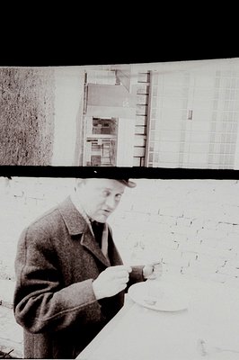 Man in a dark overcoat sits at a table, appears to be eating or drinking from a bowl. Textured brick wall backdrop with a gli...