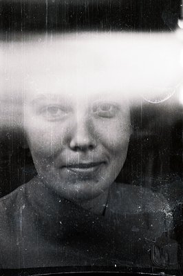 A portrait of a smiling woman, presented as a vintage photographic print. Marked by significant surface scratches and dust, s...