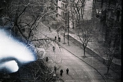 An elevated, slightly blurred view captures pedestrians walking along a lined street, flanked by bare trees. A substantial, c...