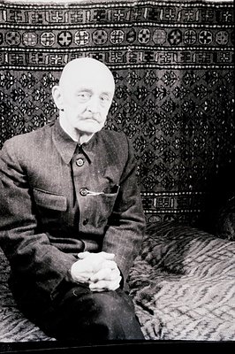 Formal portrait of an elderly man in a dark military-style suit, seated against a patterned textile backdrop. His hands are c...