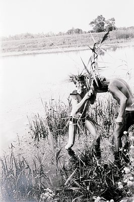 Two nude figures stand waist-deep in water amidst tall reeds and grasses along a riverbank. The male figure, positioned behin...