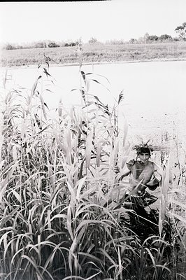 A young person, partially obscured by tall reeds, is wading in shallow water. The landscape features a wide, still body of wa...