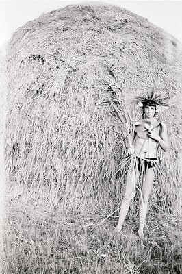 Striking black and white portrait of a shirtless male figure adorned with a headdress and grass skirt stands before a large h...