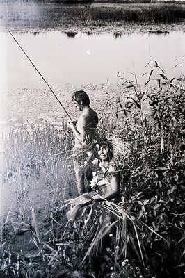 Two figures are partially concealed within tall reeds alongside a body of water. One holds a fishing rod, the other wears a f...