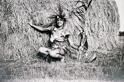 A seated figure, likely a performer or participant in a cultural ceremony, is adorned with plant material and a floral headdr...