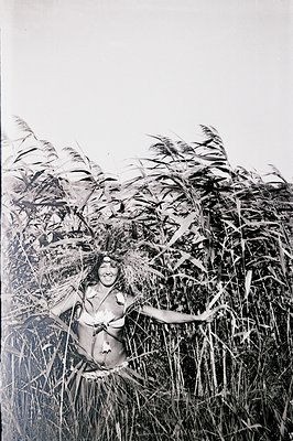 Monochrome portrait of a woman playfully posed within tall reeds or grasses. She appears to be adorned with natural foliage. ...