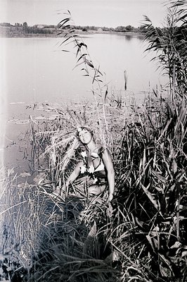 A woman reclines amidst tall reeds by a body of water, likely a lake or pond. She wears a dark swimsuit and a floral crown. T...