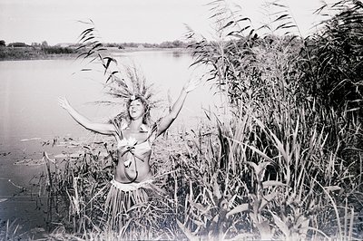 A woman with arms raised stands within tall reeds bordering a wide body of water, likely a lake or river. She wears a leafy s...
