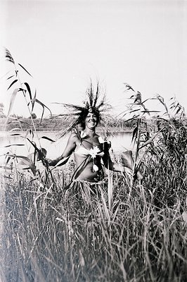 A young woman adorned with foliage and flowers stands in tall reeds beside a body of water. She wears minimal clothing, showc...