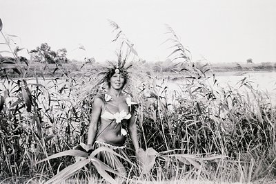 A woman adorned with elaborate headdress and skirt of natural foliage stands in tall reeds bordering a body of water. Likely ...