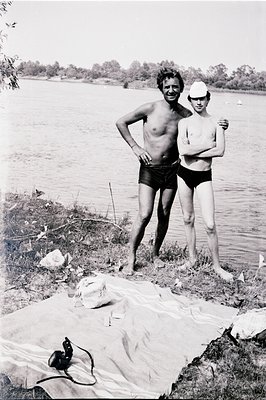 Two figures, an adult male and a young boy, stand near a waterfront, seemingly after a swim. The man wears swim trunks and a ...