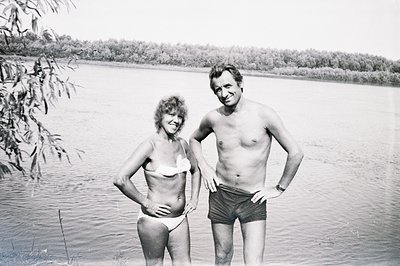 A candid, black and white snapshot captures a couple standing in shallow water near a shoreline. The woman, in a white bikini...