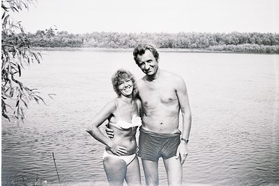 A smiling couple stands near a lakeside, the man shirtless in dark swim trunks, the woman in a white bikini. Lush foliage lin...