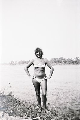 A woman in a white bikini poses on a grassy shoreline with a wide body of water behind her. Her hands are on her hips, displa...