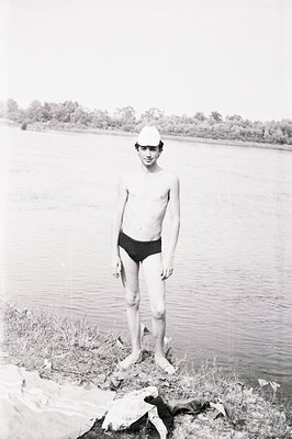 Young man in vintage swimwear stands on a grassy bank, facing the camera. A calm body of water extends into the background, f...