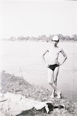 Young man stands on a grassy riverbank, dressed in swimming trunks and a swim cap. Clothing suggests a casual, recreational s...