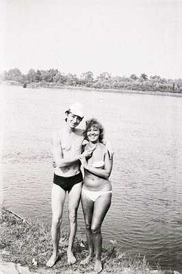 A young couple, in swimwear, stands near a body of water with dense foliage in the background. The man wears black swim trunk...
