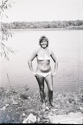 A woman in a bikini stands on a grassy shore overlooking a calm lake, hands on hips. Likely a vacation snapshot, showcasing 1...