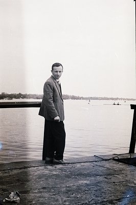 A man in a tailored suit and dark trousers stands on a weathered pier overlooking a calm, expansive body of water with distan...
