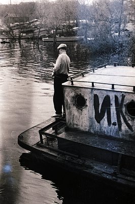 A man in a cap stands on the deck of a barge, holding a fishing rod. Cyrillic script "И.К." is painted on the vessel’s hull. ...
