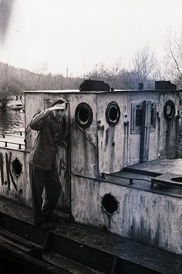 Man in a suit leans against the weathered exterior of a barge or canal boat, peering through a porthole. Industrial aesthetic...