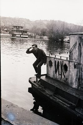 A young man in a suit poses on the edge of a stone embankment overlooking a wide, calm body of water. Cyrillic graffiti marks...