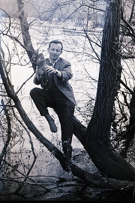 A man in a suit playfully poses perched on a fallen tree branch over a frozen lake. His confident posture and tailored clothi...