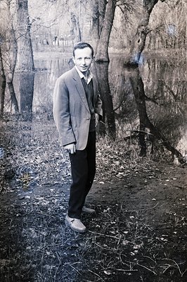 A man in a blazer and slacks stands beside a body of water, likely a pond or lake. The reflective surface mirrors the trees a...