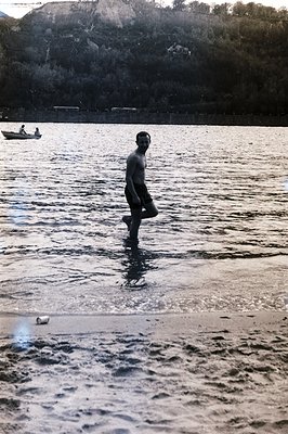 A young man wades in shallow water along a beach, possibly the Black Sea. A small rowboat with occupants is visible in the di...