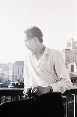 Young man in an open-collar shirt sits on a balcony overlooking a European cityscape. Architectural details suggest a possibl...