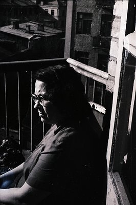 A seated woman wearing glasses gazes out from a balcony, overlooking rooftops. Strong shadows define her profile in this mood...
