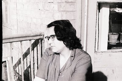 A black and white portrait captures a woman seated near a window, looking off-frame. She wears glasses and a collared shirt, ...