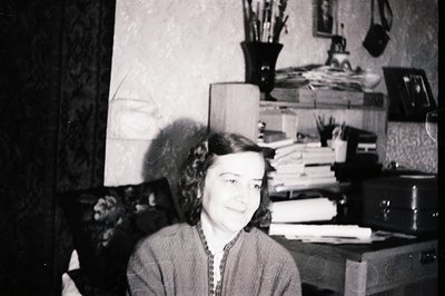 A woman sits in a cluttered office, appearing relaxed and approachable. Desk overflowing with papers and supplies hints at a ...