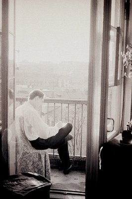 Man seated on a balcony, reading or writing. Architectural details suggest a European location, possibly Mediterranean. The s...