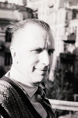 Portrait of a middle-aged man in a striped sweater and collared shirt. He appears to be outdoors, with a blurred cityscape vi...