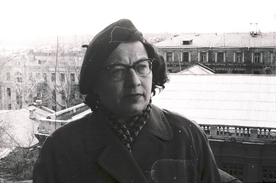 A mid-range portrait of a woman wearing glasses, a dark coat, beret, and patterned scarf. She stands against a backdrop of a ...