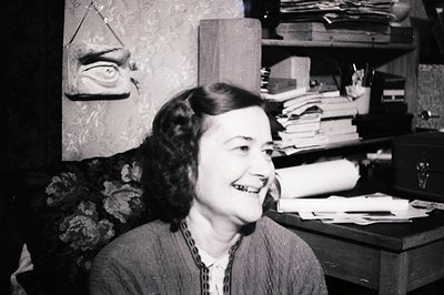 A smiling woman with shoulder-length, wavy hair sits in a richly decorated room, likely an office or study. Stacked papers an...