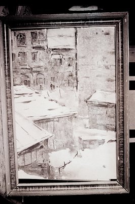 Black and white photograph depicts a snow-covered European village, likely Alpine. Sloping roofs, stone buildings, and sparse...